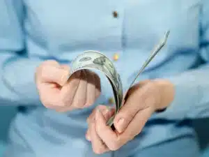 Pessoa contando notas de dólar com as mãos, vestindo camisa azul.