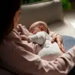 Mulher amamentando um bebê em um sofá, segurando-o com carinho enquanto ele se alimenta. Ambos estão vestidos com roupas confortáveis, e a cena transmite aconchego e cuidado maternal.