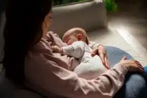 Mulher amamentando um bebê em um sofá, segurando-o com carinho enquanto ele se alimenta. Ambos estão vestidos com roupas confortáveis, e a cena transmite aconchego e cuidado maternal.
