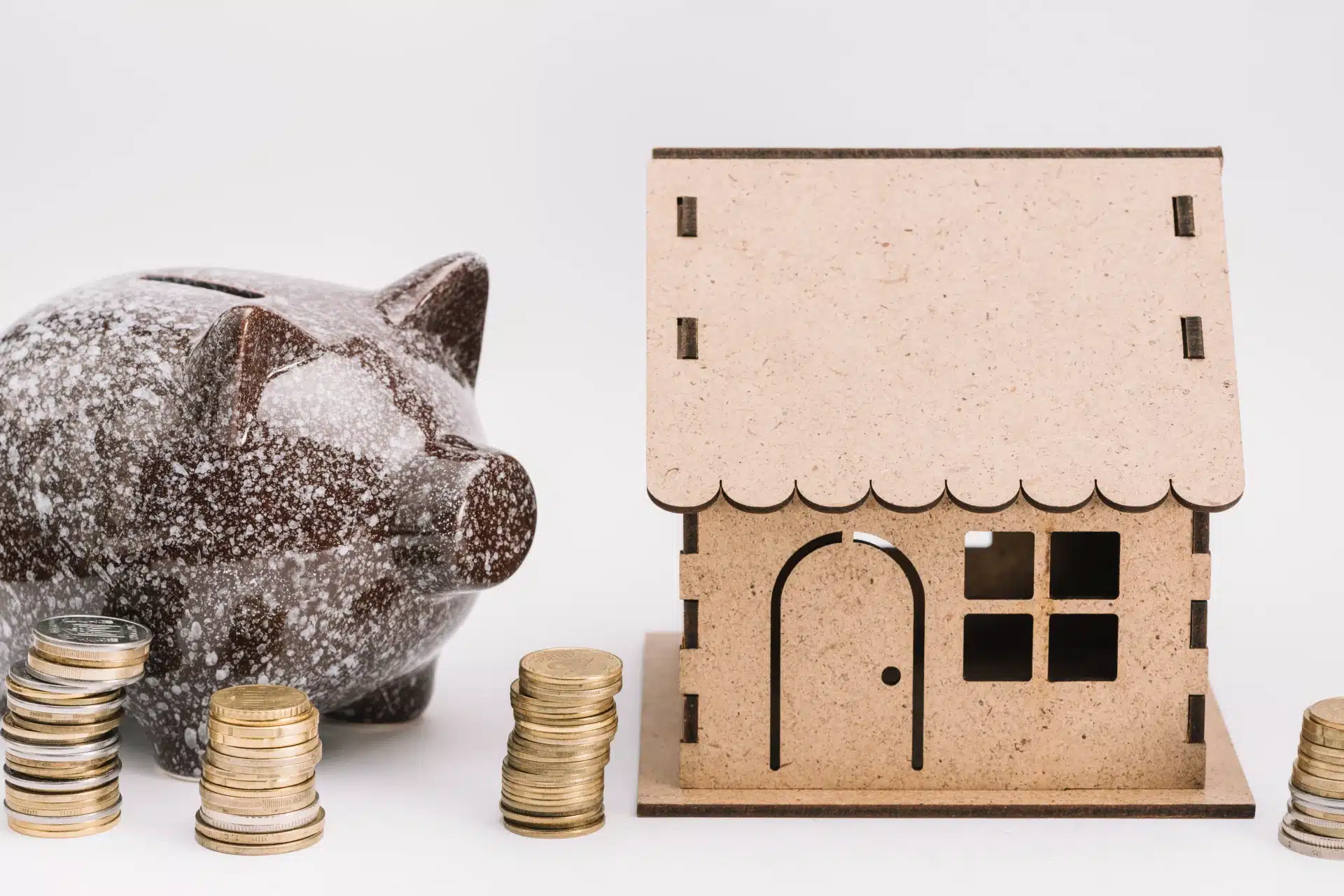 Cofrinho em formato de porco ao lado de uma miniatura de casa de madeira, com pilhas de moedas à frente, simbolizando economia e investimento em moradia.