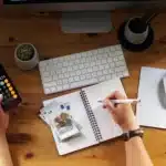 Mesa de trabalho com uma pessoa fazendo cálculos para planejamento previdenciário; uma mão segura um celular com calculadora aberta enquanto a outra anota valores em um caderno ao lado de dinheiro, documentos e um computador, representando organização financeira para o futuro.