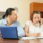 Casal sentado à mesa analisando um contrato de aluguel não renovado; a mulher fala ao telefone enquanto lê o documento com expressão preocupada, enquanto o homem ao lado observa atentamente com um laptop aberto, indicando uma situação de revisão e esclarecimento sobre o contrato.