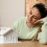 Mulher sentada à mesa observando com satisfação uma maquete de casa branca nas mãos; ao fundo há caixas de mudança, sugerindo uma situação relacionada à quitação ou baixa de hipoteca após a conclusão do financiamento imobiliário.